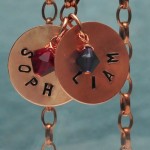 two copper discs hanging from chain sophia and a red colored birthstone hanging from one and liam and a purple birthstone hanging from second both names are stamped in a circular fashion