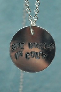 A silver disc hanging from a chain in front of a blue background it has the words she dreams in color stamped in a zigzagged line across it